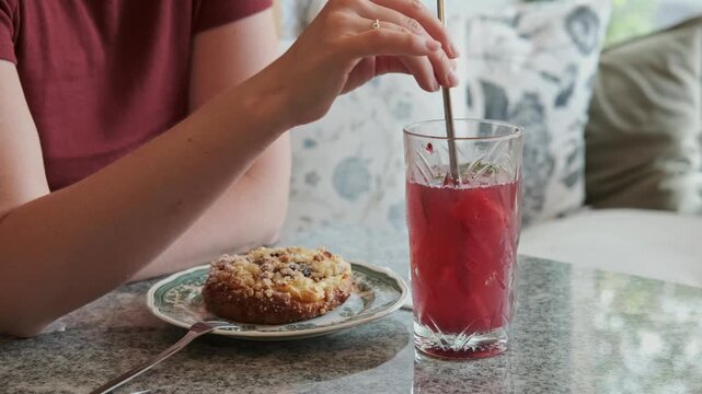 Female hand stirs a refreshing red beverage with a straw while sitting at a table with a plate of dessert in a cozy indoor setting with patterned cushions