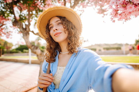 Close-up of a young woman taking a selfie with her phone on a sunny day near flowers. The young woman blogs and enjoys the outdoors. Concept of beauty, blogging, and enjoyment.