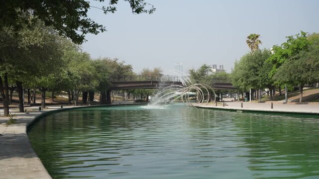 A peaceful view of the Santa Luc&iacute;a Riverwalk passing beneath a bridge at Fundidora Park in Monterrey, Nuevo Le&oacute;n, Mexico. Crystal-clear water, decorative fountains, and lush green surroundings create 