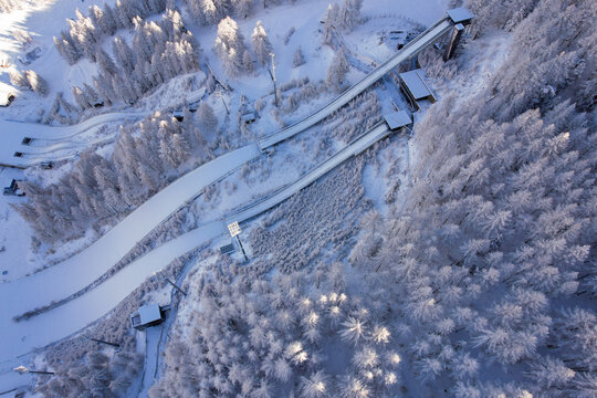 Aerial view of Stadio Olimpico del Salto ski jumping facility surrounded by snow-covered pine forests in the mountains of Pragelato, Piedmont, Italy.
