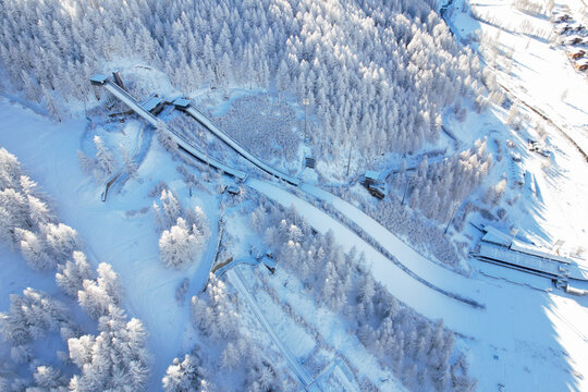 Aerial view of Stadio Olimpico del Salto ski jumping facility surrounded by snow-covered pine forests and mountain landscape in Pragelato, Piedmont, Italy.