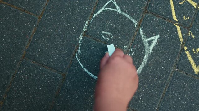 Child drawing a cat face with blue chalk on dark street pavement. Creative hobby of a kid playing outdoors. Artistic activity on concrete for recreation concept.