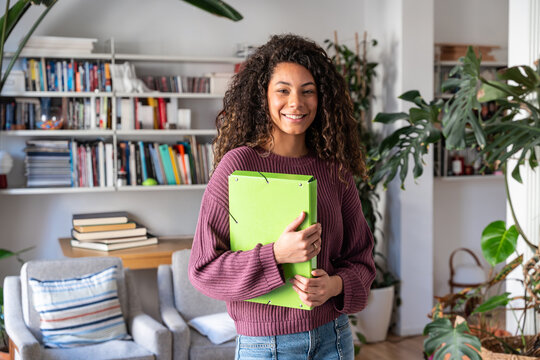 Smiling latin student holding folder in home study room