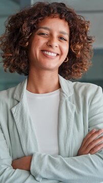 Vertical portrait of smiling young multiracial latin or indian businesswoman with curly hair standing in office, wearing blazer in professional workspace. Arabian young woman girl smiling at camera