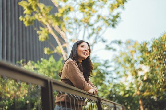 Happy young Asian woman in a brown sweater and white shirt smiling while leaning on a balcony railing in a green park during golden hour. A peaceful female professional enjoying a break outdoors.