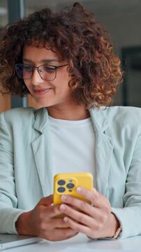 Young curly hair Indian business woman manager using cell phone mobile app. Smiling Latin Hispanic mature female businesswoman holding smartphone in office trading, reading, working online. Vertical