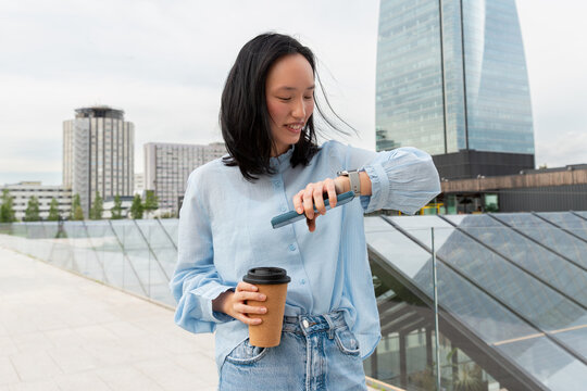 Gen Z Asian woman checking smartwatch with coffee outdoors