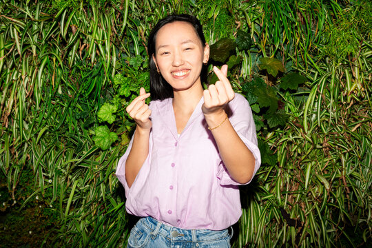 Gen Z Asian woman making finger heart gesture by greenery