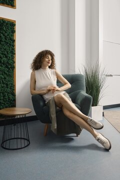Woman relaxing in a stylish chair in a modern office space with greenery and natural light. She has curly hair and wears a casual outfit suitable for everyday wear