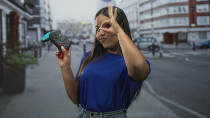 Young woman holding a gamepad while smiling and making an ok sign over her eye on a street with blurred buildings and parked cars  playful fun. © Krakenimages.com