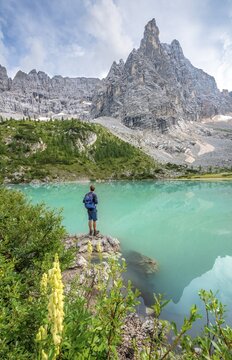 Young man hiker at the turquoise-green Sorapis lake with flowers, Lago di Sorapis, mountain peak Dito di Dio, Dolomites, Belluno, Italy