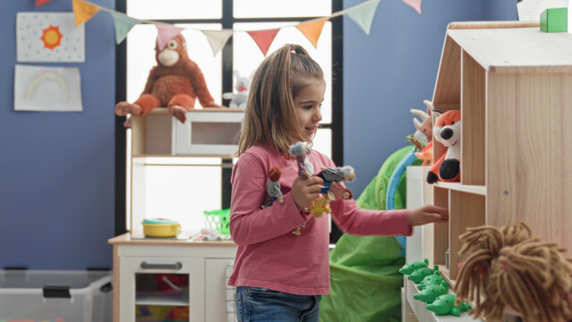 Girl holding several dolls and reaching for a small toy on a wooden shelf in a colorful playroom inside a building; playful curiosity.