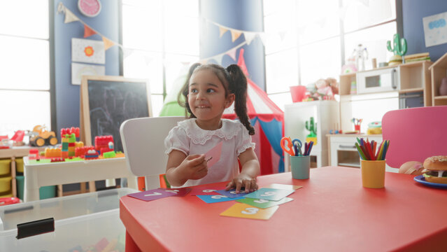 Young girl sorting colorful number cards with small hands at a red table in a building playroom with toys and craft cups; concentration learning play.