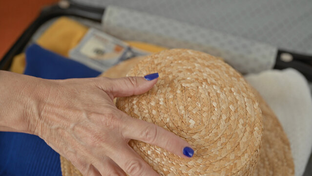Woman packing a hard shell suitcase, hand with blue nails holding a straw hat and visible dollars on bed in building; anticipation planning.