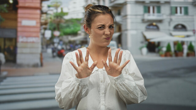 Young woman in white blouse grimacing with clasped hands on a busy city street crosswalk near cafe storefronts; disgust.