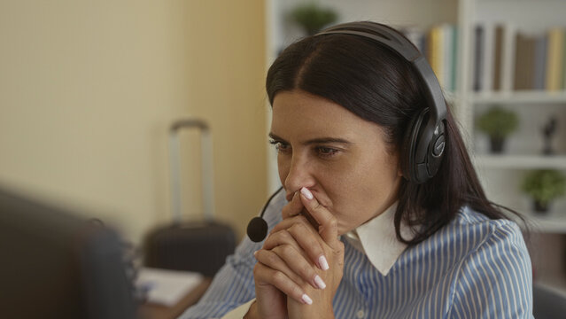 Woman with headset pressing finger to lips and clasping hands at computer desk in building; concern problem solving.