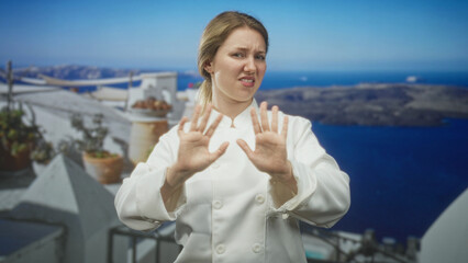Fototapeta premium Woman chef in white uniform holds hands up palms forward on sunlit balcony overlooking sea and white buildings; disgust rejection aversion.