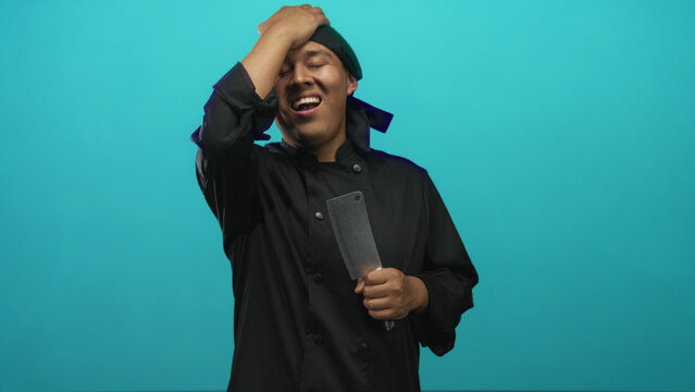 Man chef in black uniform holding a cleaver while pressing his forehead with one hand in a turquoise studio; kitchen exhaustion.