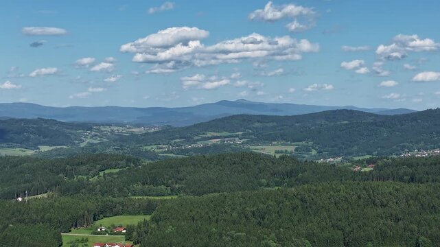 Die Kamera zeigt eine weite Landschaft mit dem Gro&szlig;en Arber im Hintergrund. Wolken ziehen &uuml;ber den Himmel. B&auml;ume und Wiesen sind sichtbar. Es ist ein klarer Tag.