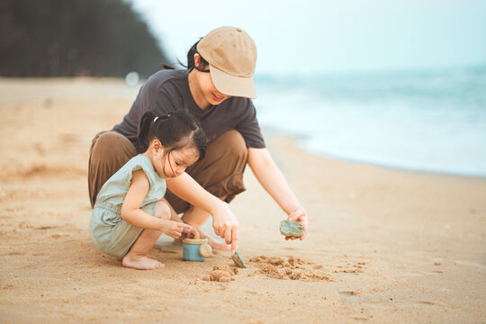 Asian mother and little daughter crouching and playing with sand toys on beach, happy family building sandcastles during summer holiday vacation for travel tourism, parenting.