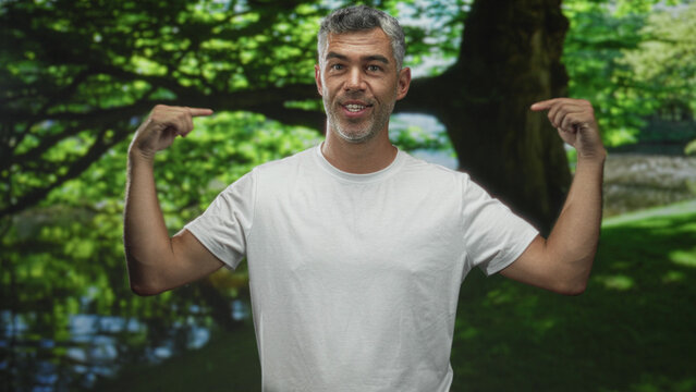 Man in white tshirt points fingers to chest with smirk, bare forearms visible, standing by large tree and pond in forest; confidence self.