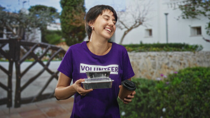 Obraz premium Woman volunteer holding foil food containers and a coffee cup, smiling while standing in front of a building; community care compassion.