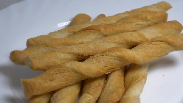 Close-up of crispy twisted puff pastry breadsticks rotating on a white plate, showcasing their golden flaky texture under bright studio lighting.