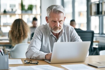 Fototapeta premium Senior man works on laptop at a desk in a modern office space during the day with other people present in the background