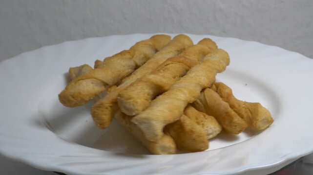 Close-up of crispy twisted puff pastry breadsticks rotating on a white plate, showcasing their golden flaky texture under bright studio lighting.