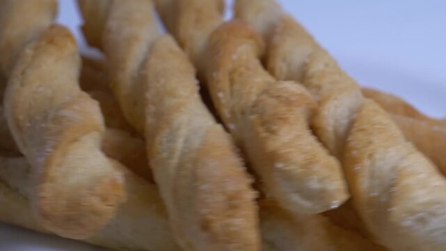 Close-up of crispy twisted puff pastry breadsticks rotating on a white plate, showcasing their golden flaky texture under bright studio lighting.