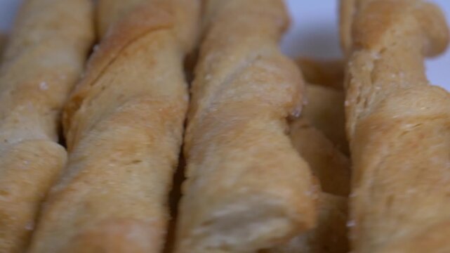 Close-up of crispy twisted puff pastry breadsticks rotating on a white plate, showcasing their golden flaky texture under bright studio lighting.