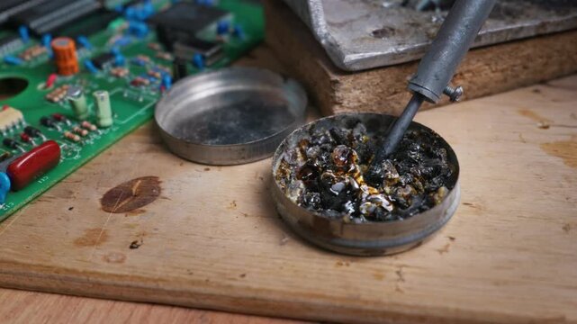 A hot soldering iron tip melts dark rosin flux inside a metal tin on a wooden workbench next to a printed circuit board. Concept of electronic repair, engineering, and maintenance.