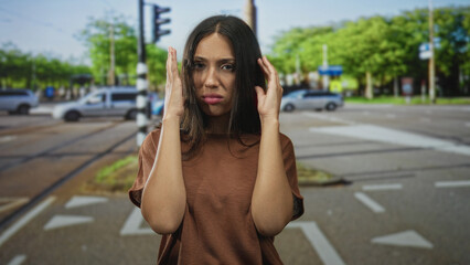 Fototapeta premium Hispanic woman pressing hands to ears at a busy street intersection with traffic and tram tracks; traffic noise annoyance.