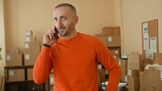 Man in orange sweater holding smartphone to ear beside stacked cardboard boxes in a building; determination small business.