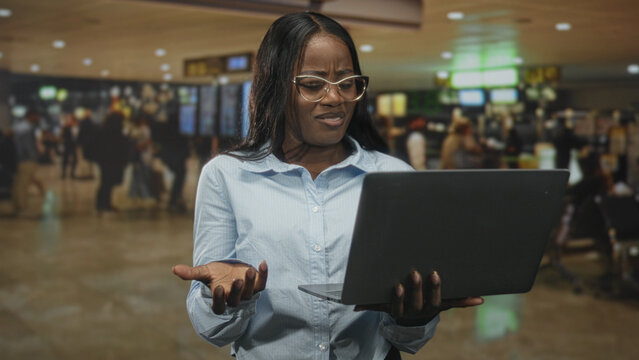 Woman holding laptop, typing and frowning while checking screen near departure boards in airport terminal; frustration problem solving.