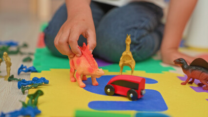 Boy kneeling on foam playroom mat arranging toy dinosaurs and a red toy car with hand reaching  imaginative play joy. © Krakenimages.com