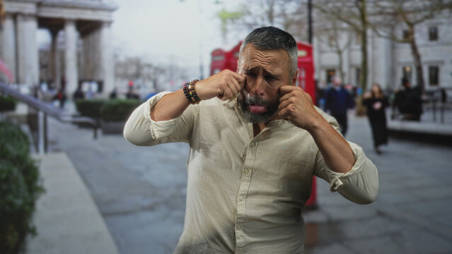 Middle aged hispanic man with gray beard rubs eyes and pulls a funny face on street near a red telephone kiosk by stone building; playfulness.