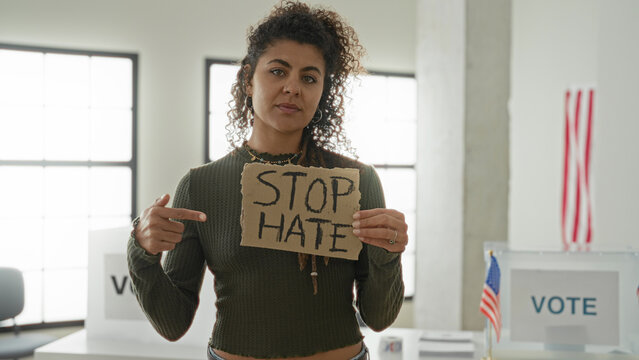 Woman holding cardboard sign reading stop hate while showing palm out toward camera inside a voting building with vote booths and us flags visible; solidarity protest.
