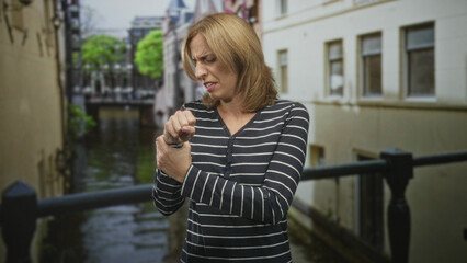 Fototapeta premium Woman clutching wrist in pain beside building railing on a canal walkway, holding a watch and grimacing; pain concern.