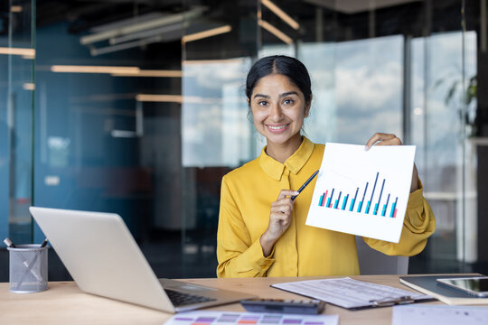 Portrait of a smiling and successful Indian woman in the office, sitting at the desk and pointing at the camera with a pen