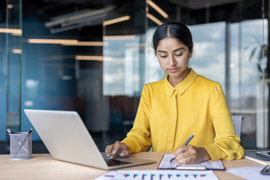 A young Indian businesswoman is busy taking notes in a document and typing on a laptop in the office