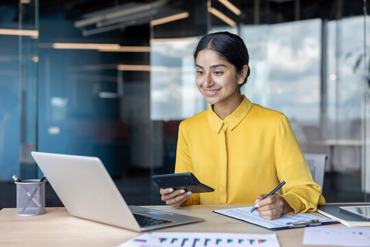 Smiling young Indian businesswoman working in the office at a desk with a laptop, taking notes and using a calculator