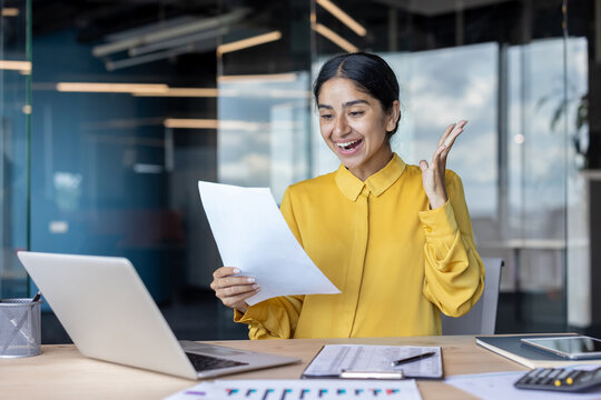 Happy Indian young woman sitting at office desk and rejoicing, reading document and agreement which she holds in her hand