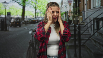 Fototapeta premium Young woman in red plaid shirt with hands raised and wide eyes, standing by a bicycle on a tree lined street; surprise explanation.