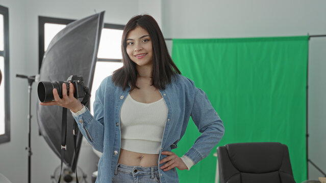 Young woman holding camera with hand on hip in a studio with green screen and lighting; confidence creativity.