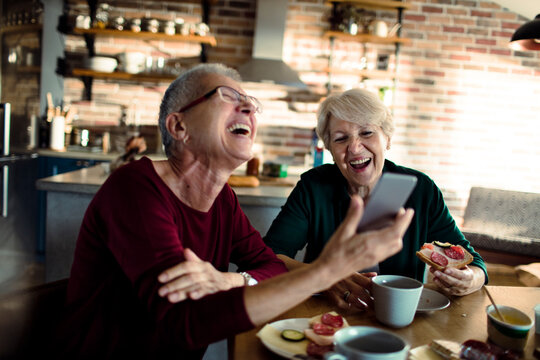 Two senior women laughing over a smartphone in a home kitchen