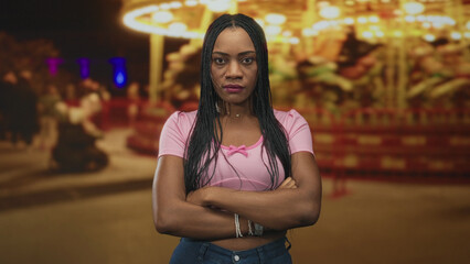 Fototapeta premium Woman with arms crossed and exposed forearms standing by a carousel on a street fair at night, looking sideways and unimpressed; defiance solitude.