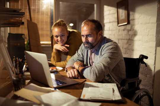 Man in wheelchair working on laptop with woman at home office at night