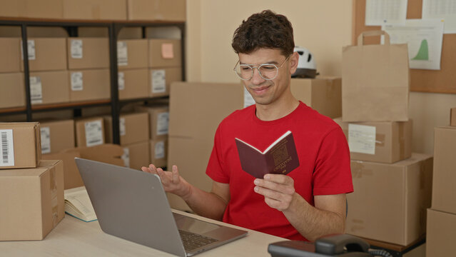 Man holding passport and reading laptop while packing parcels at a small business building; logistics planning concentration.