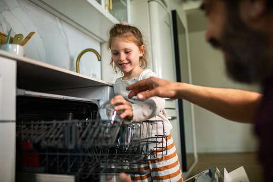 Smiling girl helping parent load dishwasher at home kitchen